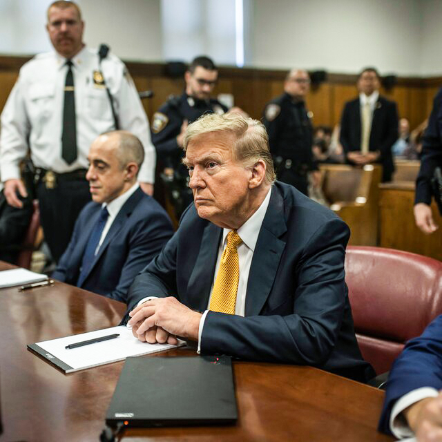 Donald Trump at the defense table in a navy suit.