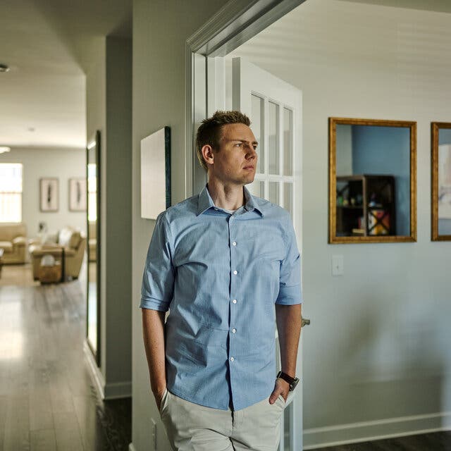 Travis Wattles is standing inside his living room, wearing a blue shirt. 